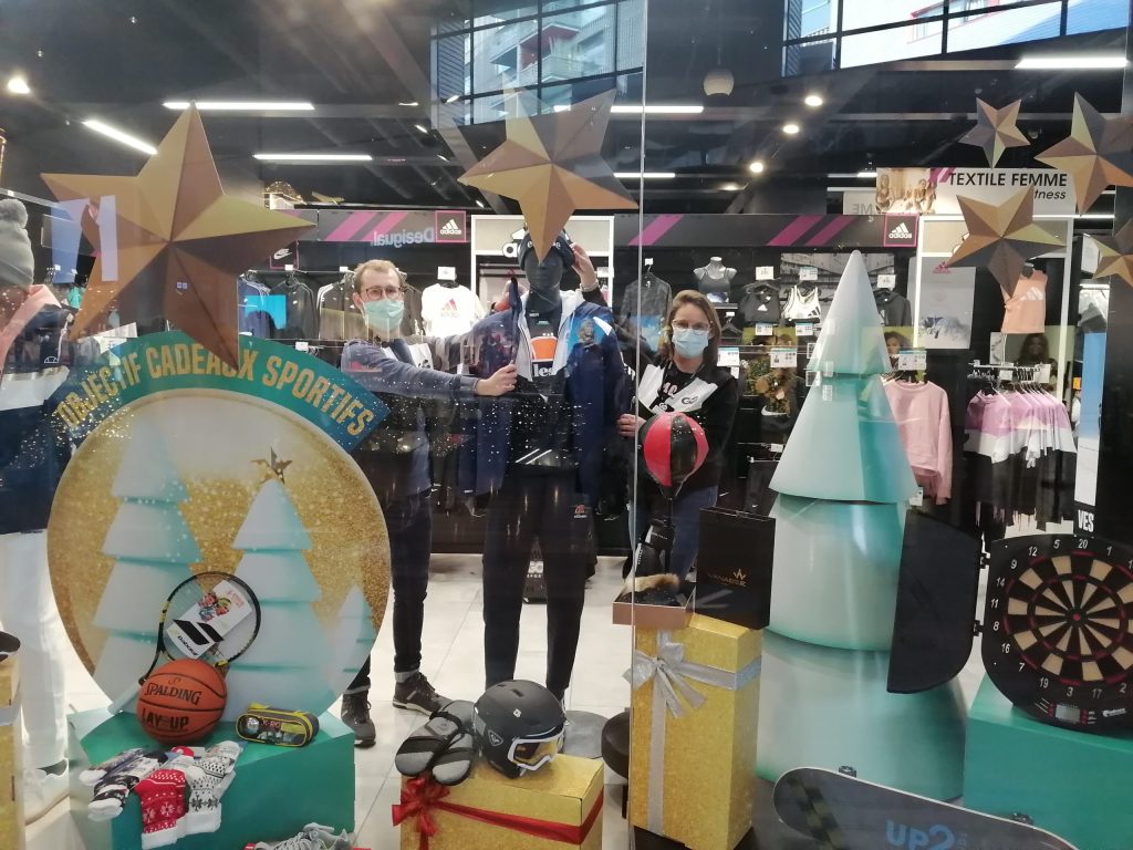 Deux personnes masquées derrière une vitrine de Noël de magasin de sport, banderole « Objectif cadeaux sportifs ».