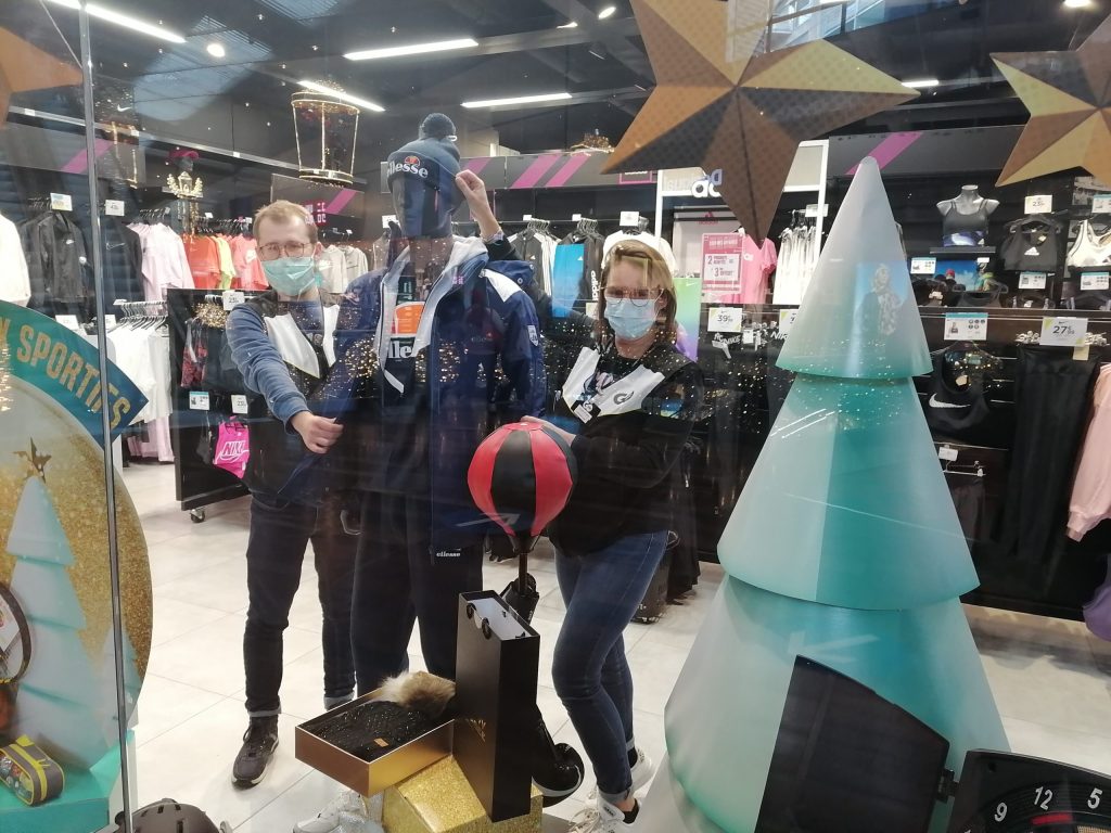 Deux personnes masquées installent des décorations de Noël dans la vitrine d'un magasin de sport.