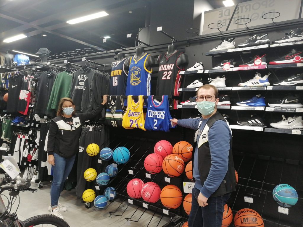 Deux personnes masquées devant le rayon basket d'un magasin de sport, maillots NBA et ballons exposés.