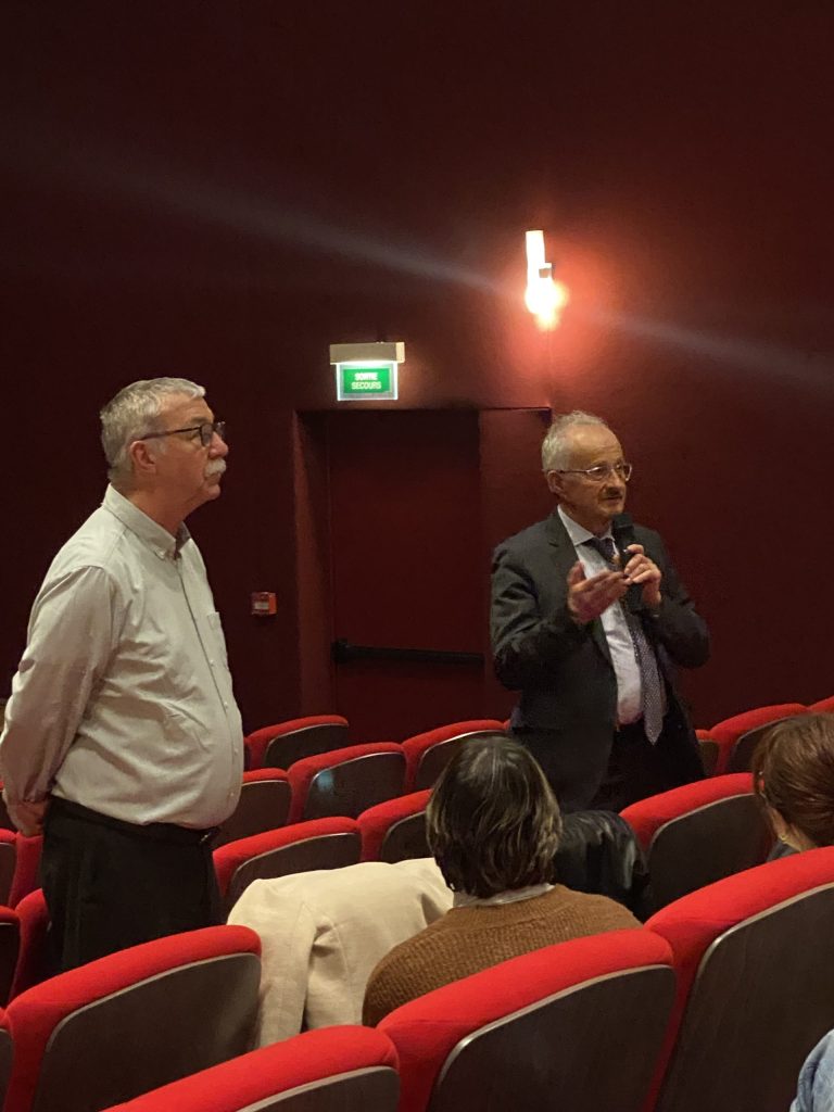 Deux hommes debout dans une salle de cinéma aux fauteuils rouges, l'un s'adressant au public au micro.