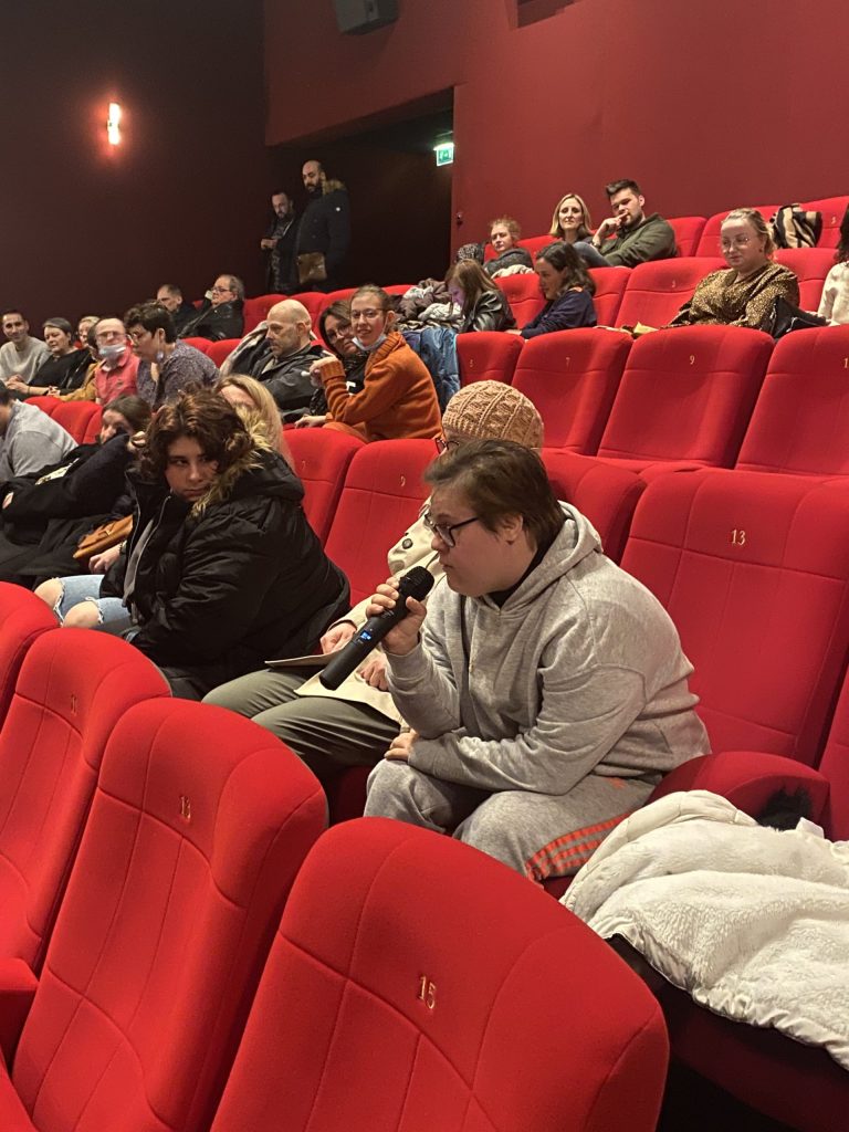 Dans une salle aux fauteuils rouges, une spectatrice prend la parole au micro devant le public assis.