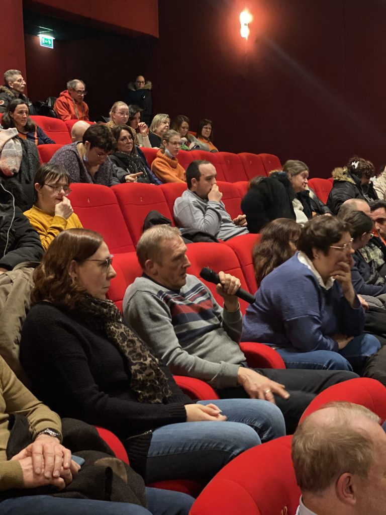 Public installé dans les fauteuils rouges d'une salle de spectacle ; un spectateur tient un micro pour poser une question.