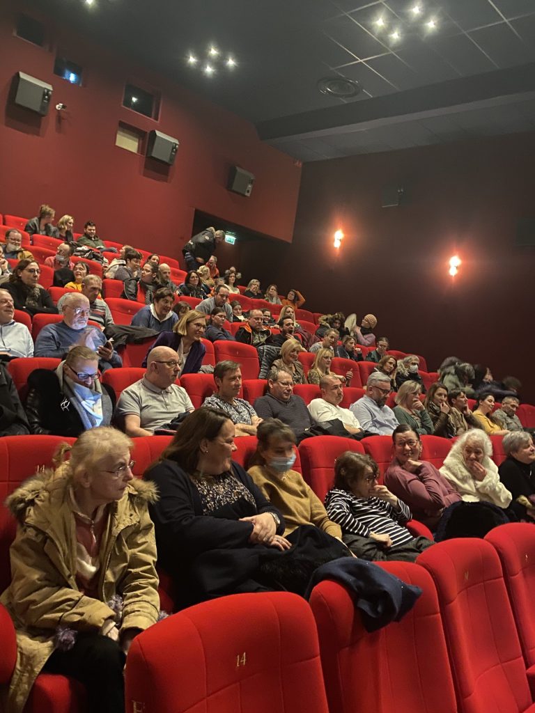 Public assis dans les fauteuils rouges d'une salle de cinéma, la plupart tournés vers l'écran.
