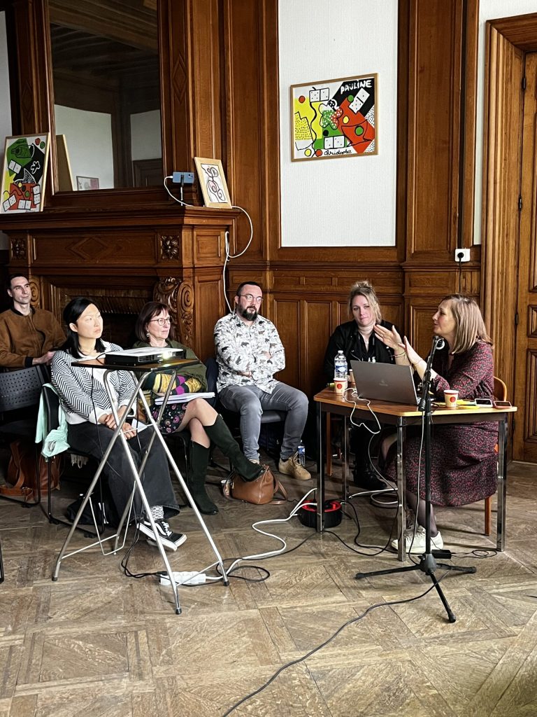 Dans une salle lambrissée, une intervenante prend la parole au micro devant un petit groupe assis qui l'écoute.