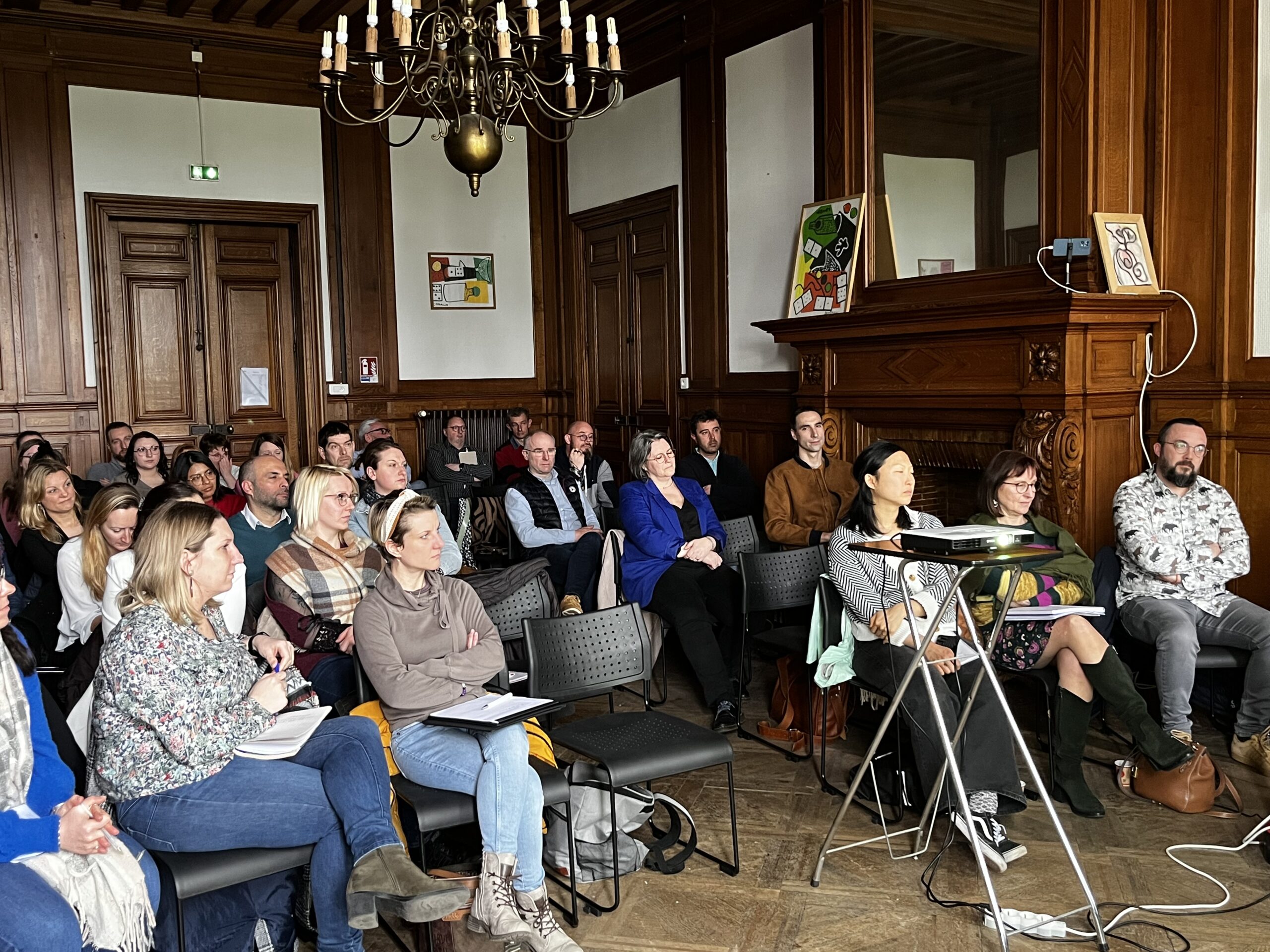 Un public nombreux assiste à une présentation dans une salle lambrissée, projecteur sur trépied
