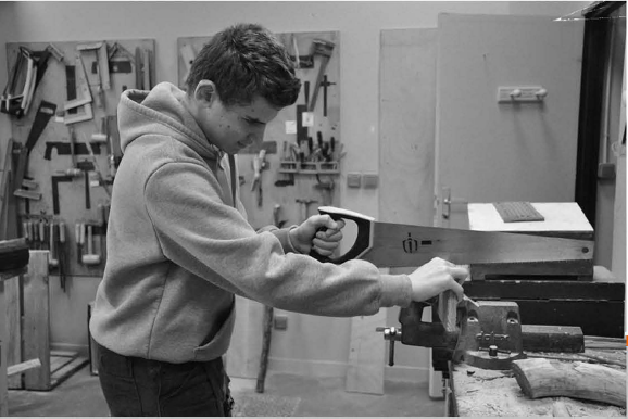Un adolescent scie une planche serrée dans un étau, au milieu des outils d'un atelier de menuiserie