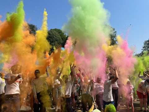 Des participants en t-shirt blanc lancent des poudres colorées, bras levés sous un ciel bleu