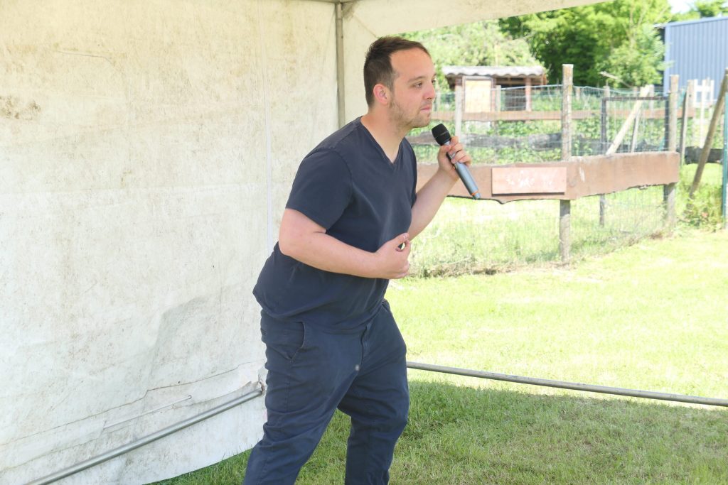 Sous un chapiteau, un homme s'adresse au public avec un micro, un enclos et un jardin en arrière-plan.
