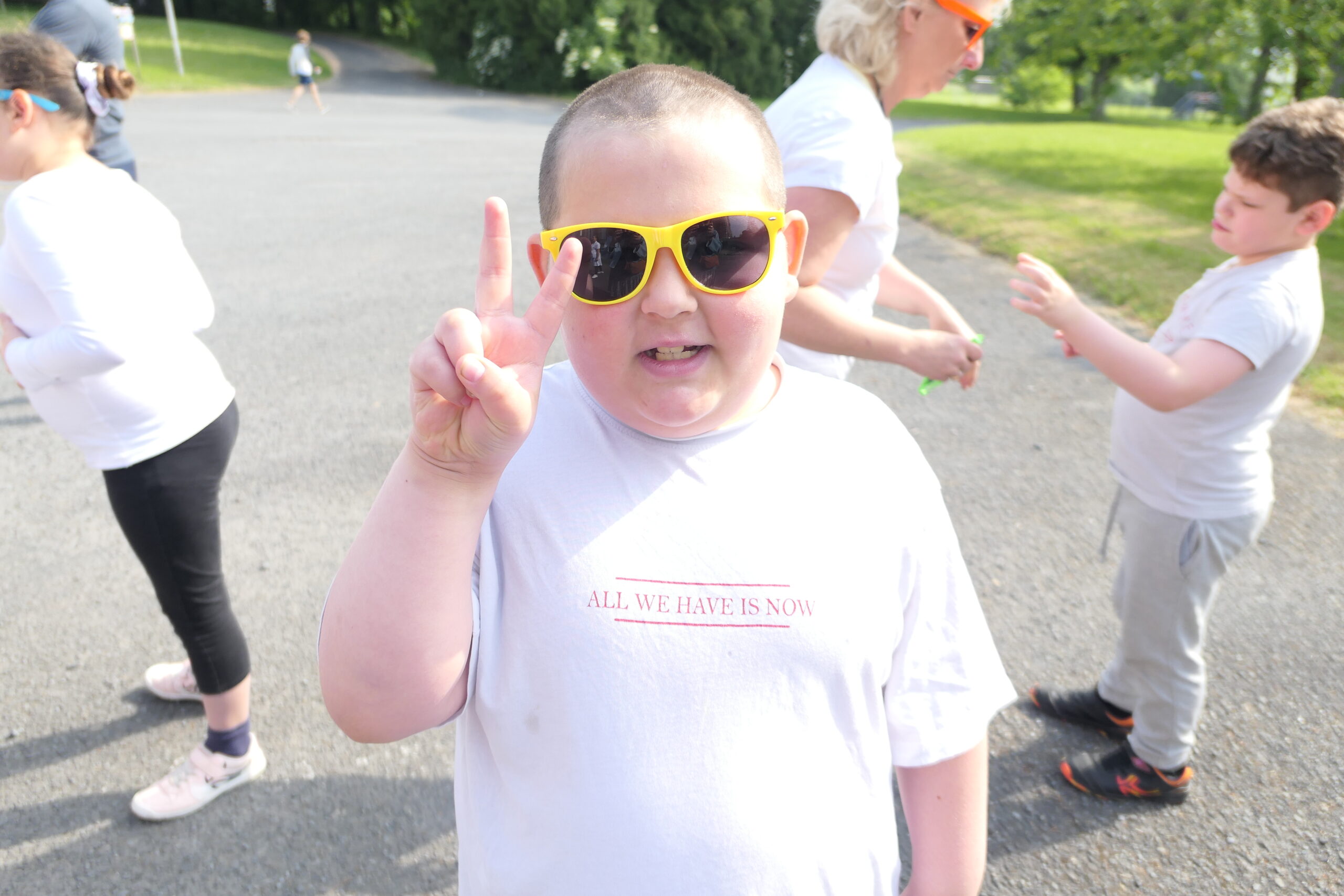 Un enfant portant des lunettes de soleil jaunes fait le signe V pendant une marche collective en plein air.