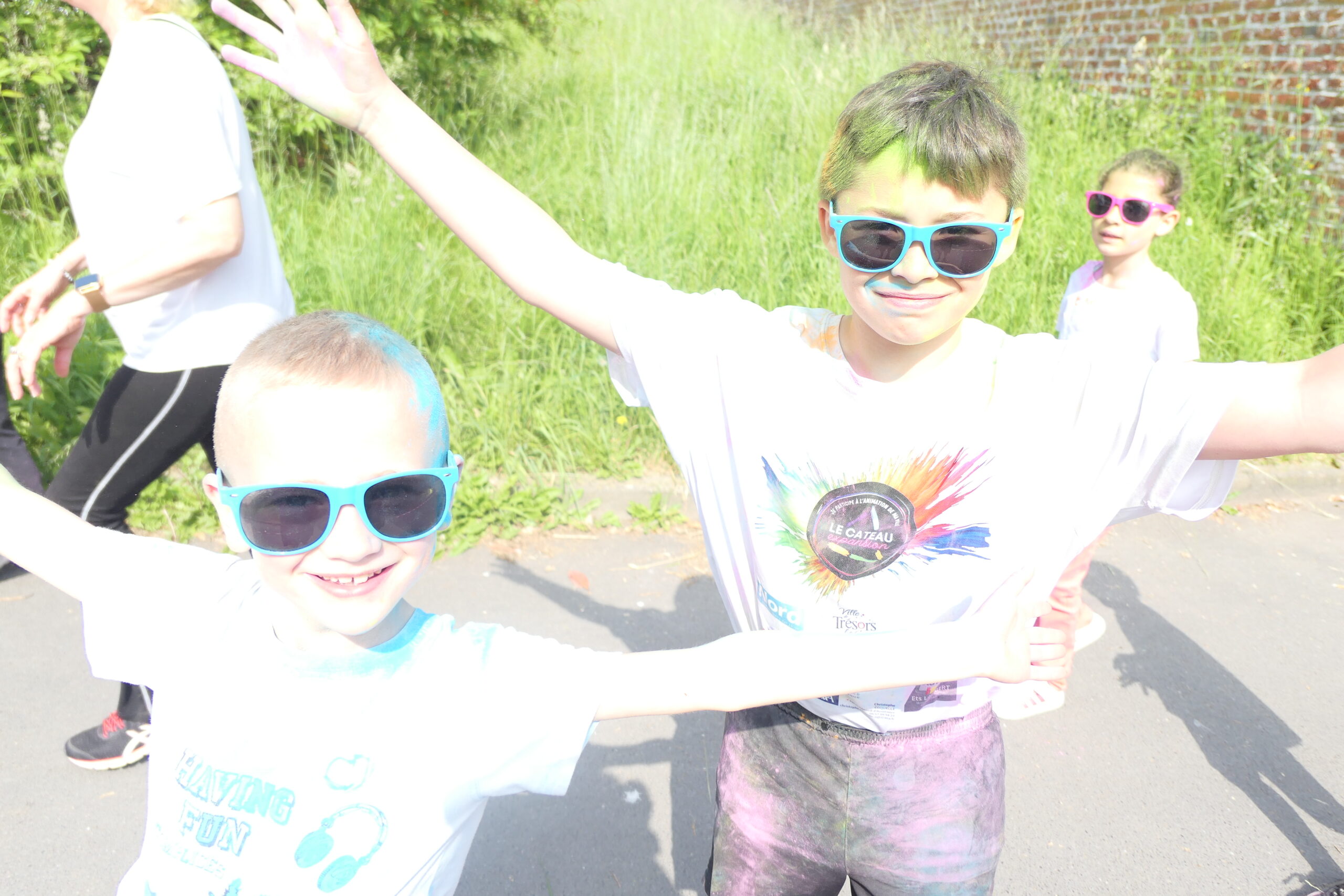 Deux enfants en lunettes de soleil, cheveux et vêtements couverts de poudre colorée, bras écartés sur la route.
