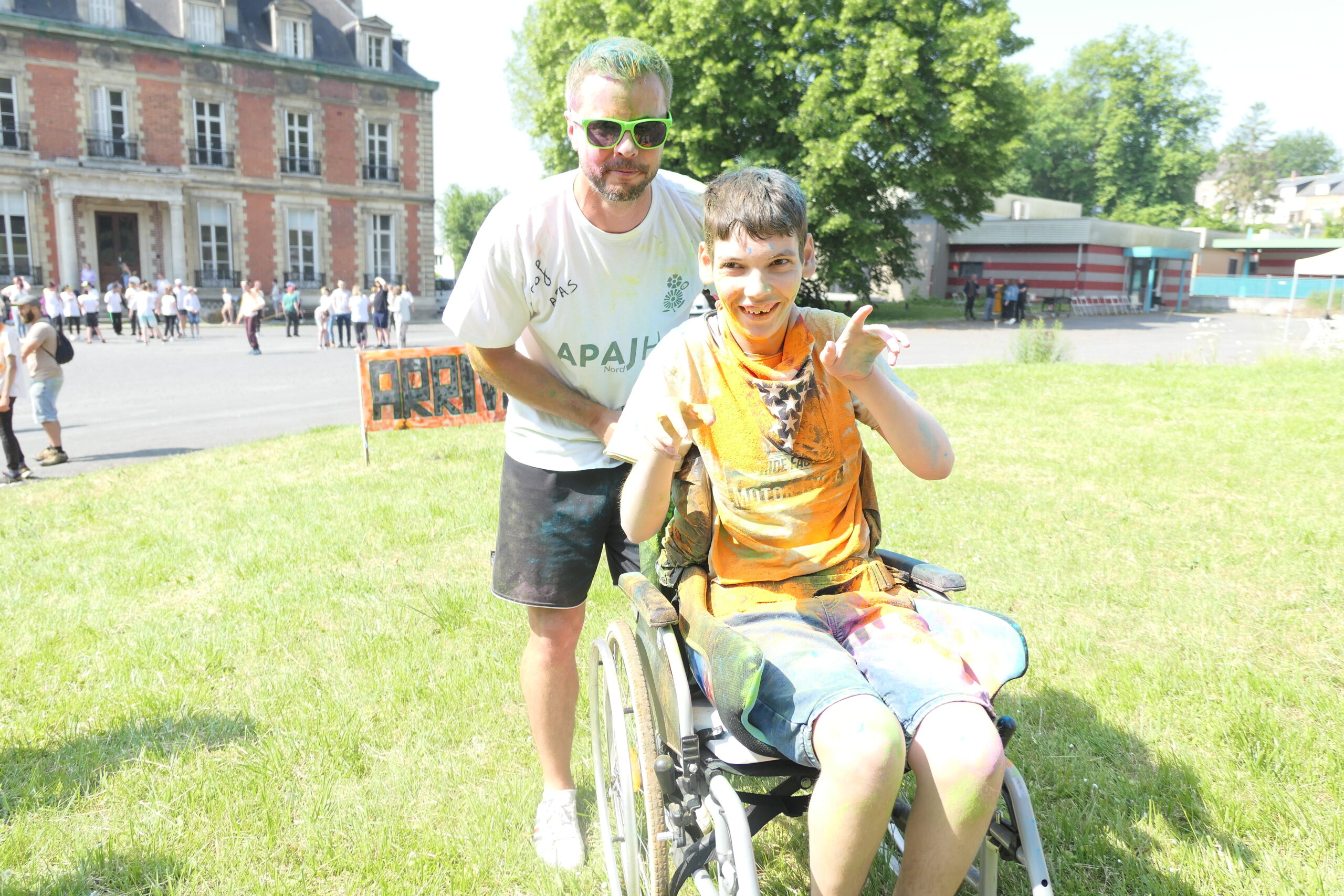 Couverts de poudre colorée, un homme en tee-shirt APAJH Nord pousse un jeune en fauteuil roulant sur la pelouse.