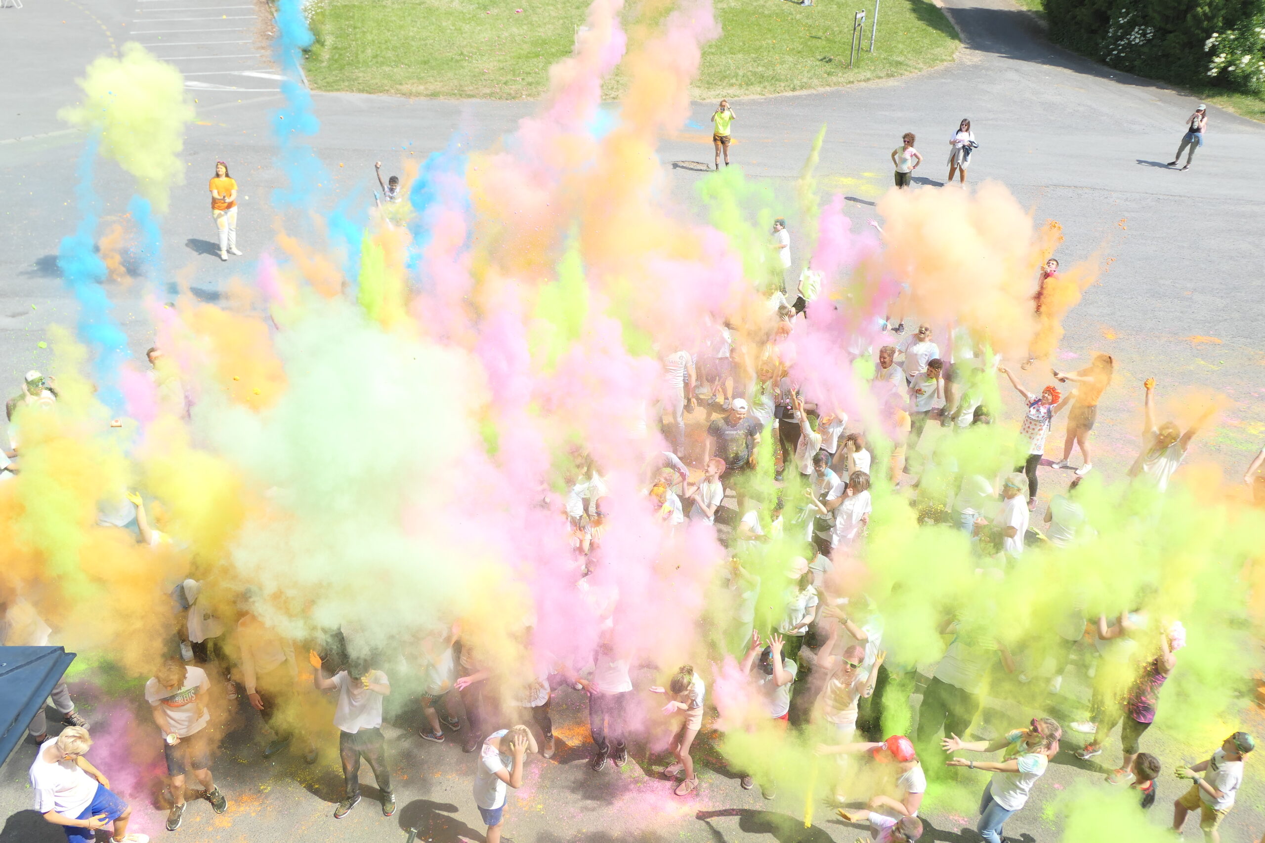 Vue en plongée d'une foule vêtue de blanc lançant des poudres colorées lors d'une color run sur un parking.