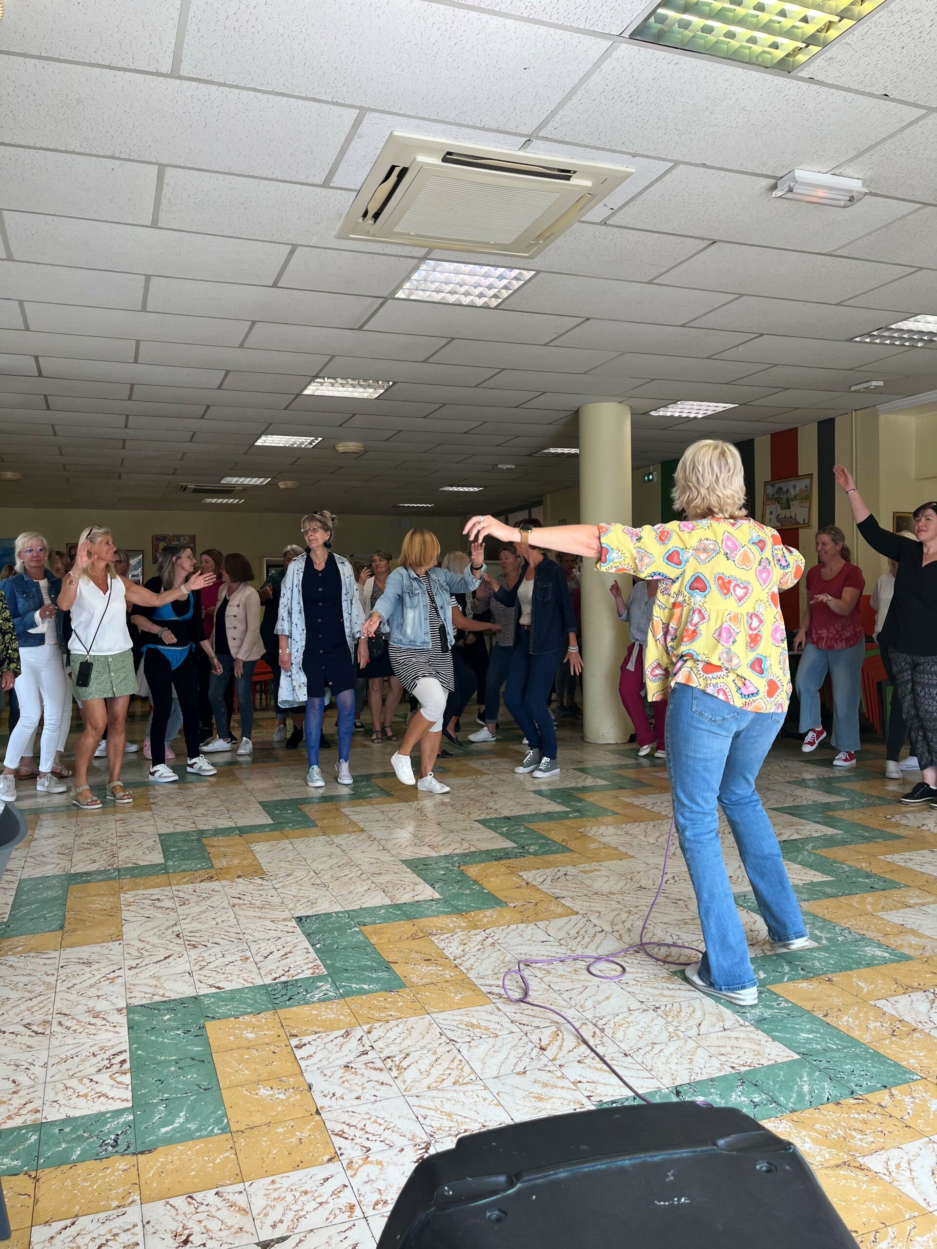 Une animatrice guide une salle remplie de participantes qui reprennent ses mouvements de danse.