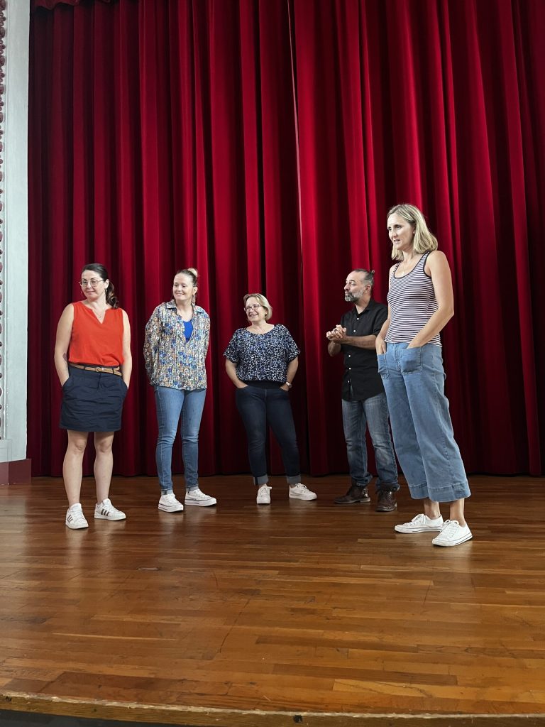 Cinq personnes debout sur la scène d'une salle de spectacle, devant un rideau rouge ; l'une s'adresse au public.