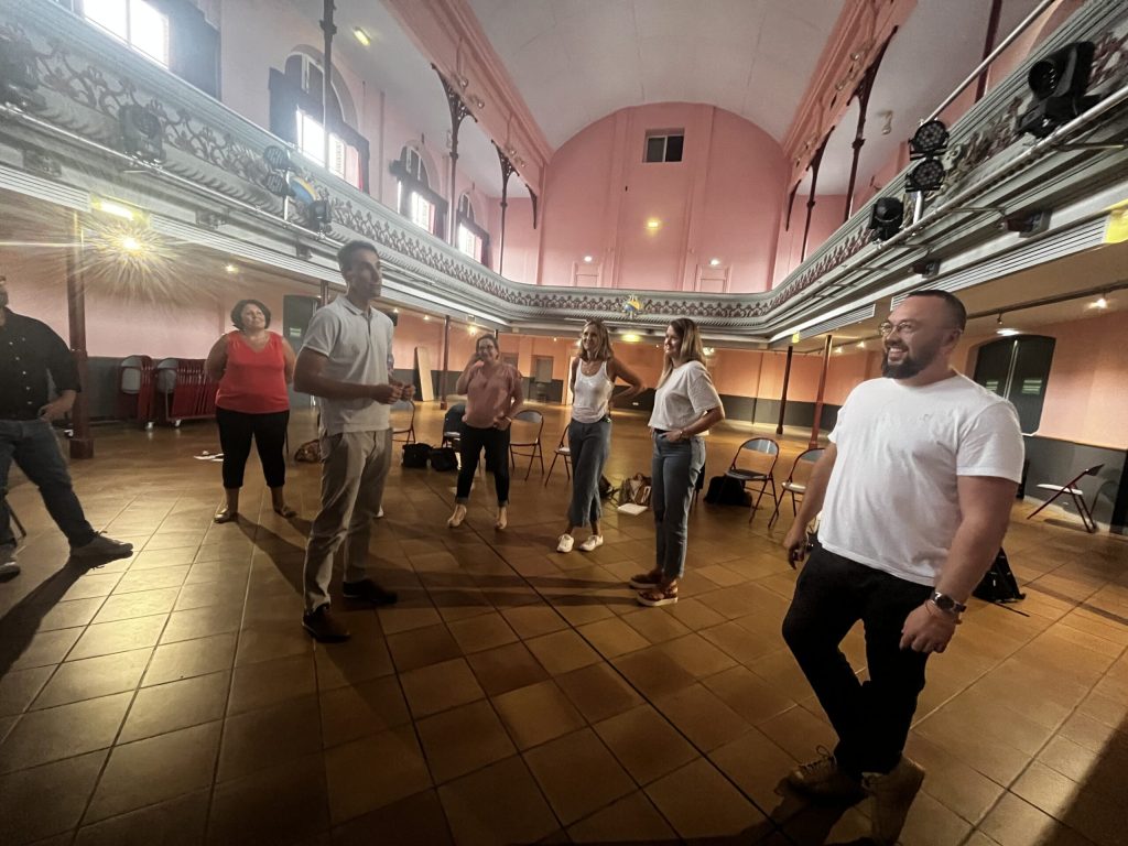 Plusieurs personnes debout, espacées, dans une grande salle des fêtes, lors d'un atelier collectif.
