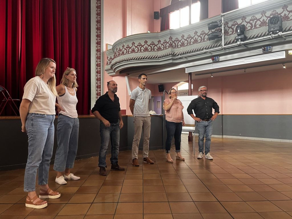 Six personnes debout face au public dans une salle de spectacle, rideau rouge et balcon à l'arrière.