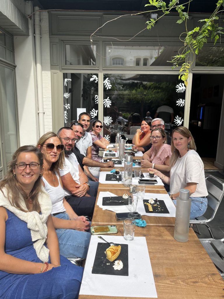 Une dizaine d'adultes attablés en terrasse posent en souriant à la fin d'un repas, desserts encore servis.