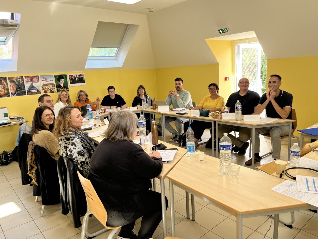 Une quinzaine de personnes réunies autour de tables en U dans une salle de réunion aux murs jaunes.