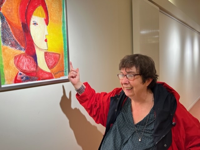 Une femme souriante désigne du doigt un portrait peint aux couleurs vives accroché au mur.