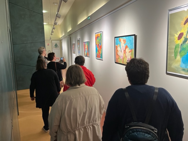Des visiteurs longent un couloir où sont accrochées des peintures colorées, vaches et bouquets de tournesols.