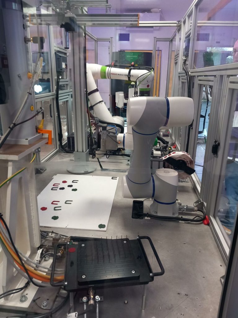 Bras robotisé collaboratif dans une cellule vitrée d'atelier, devant un plateau de pièces à assembler.