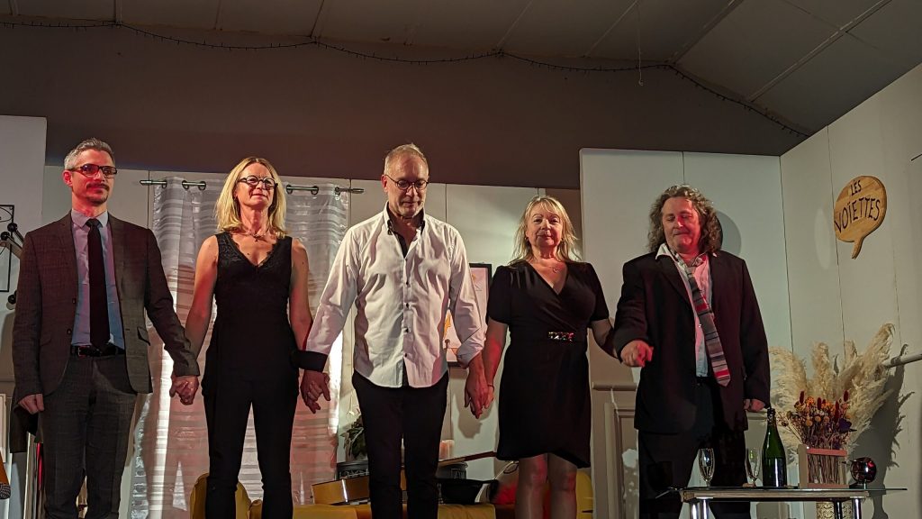 Cinq comédiens saluent main dans la main sur scène, devant un panneau en forme de bulle « Les Noisettes ».