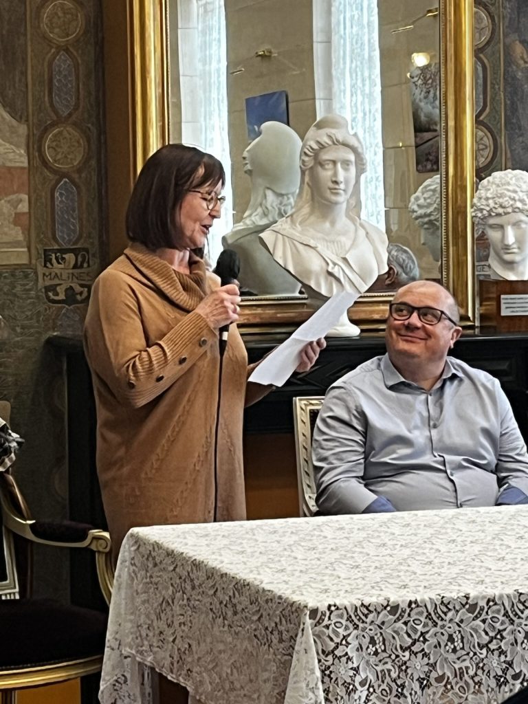 Une femme lit un texte au micro dans un salon orné de bustes et de miroirs dorés ; un homme assis l'écoute.
