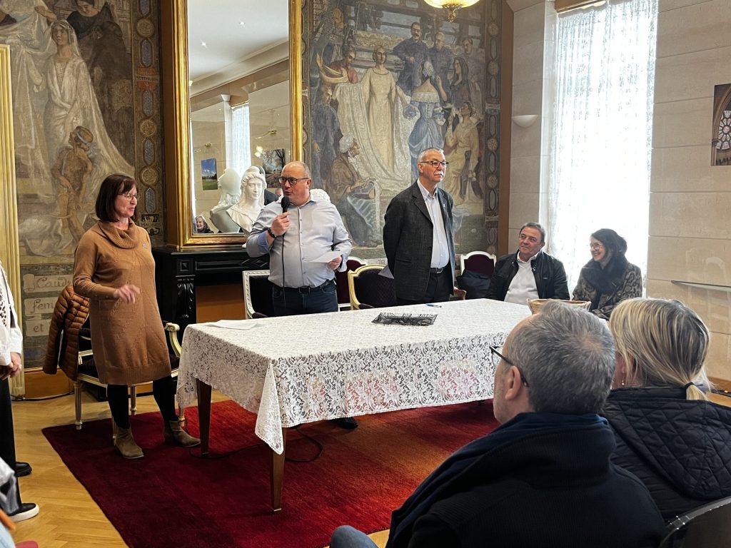 Un homme s'exprime au micro devant un public, entouré de plusieurs personnes, dans un salon orné de fresques.
