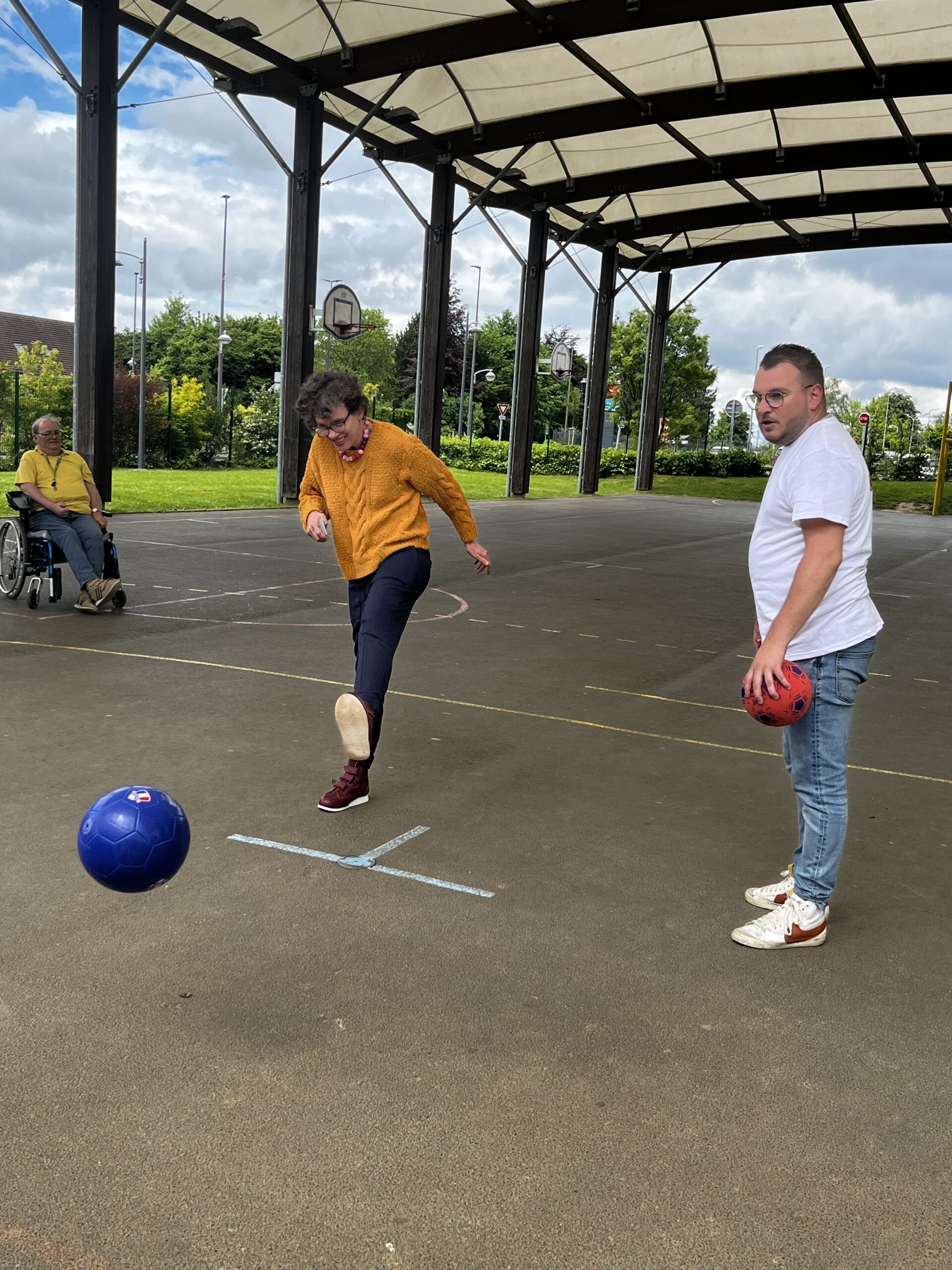 Sur un terrain de sport couvert, une femme frappe un ballon du pied face à un homme qui en tient un autre.