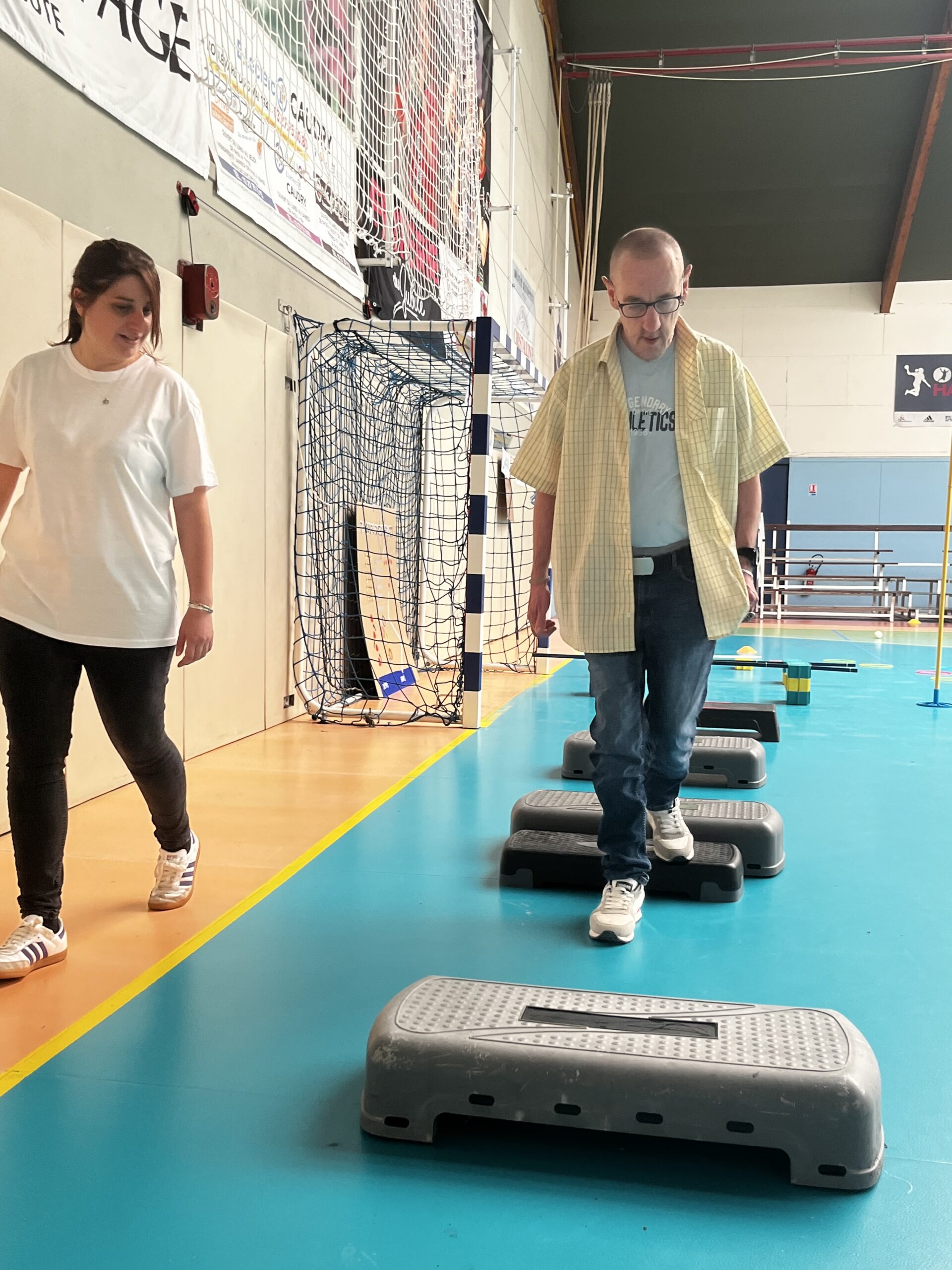 Dans un gymnase, un homme franchit un parcours de steps sous le regard d'une accompagnante.