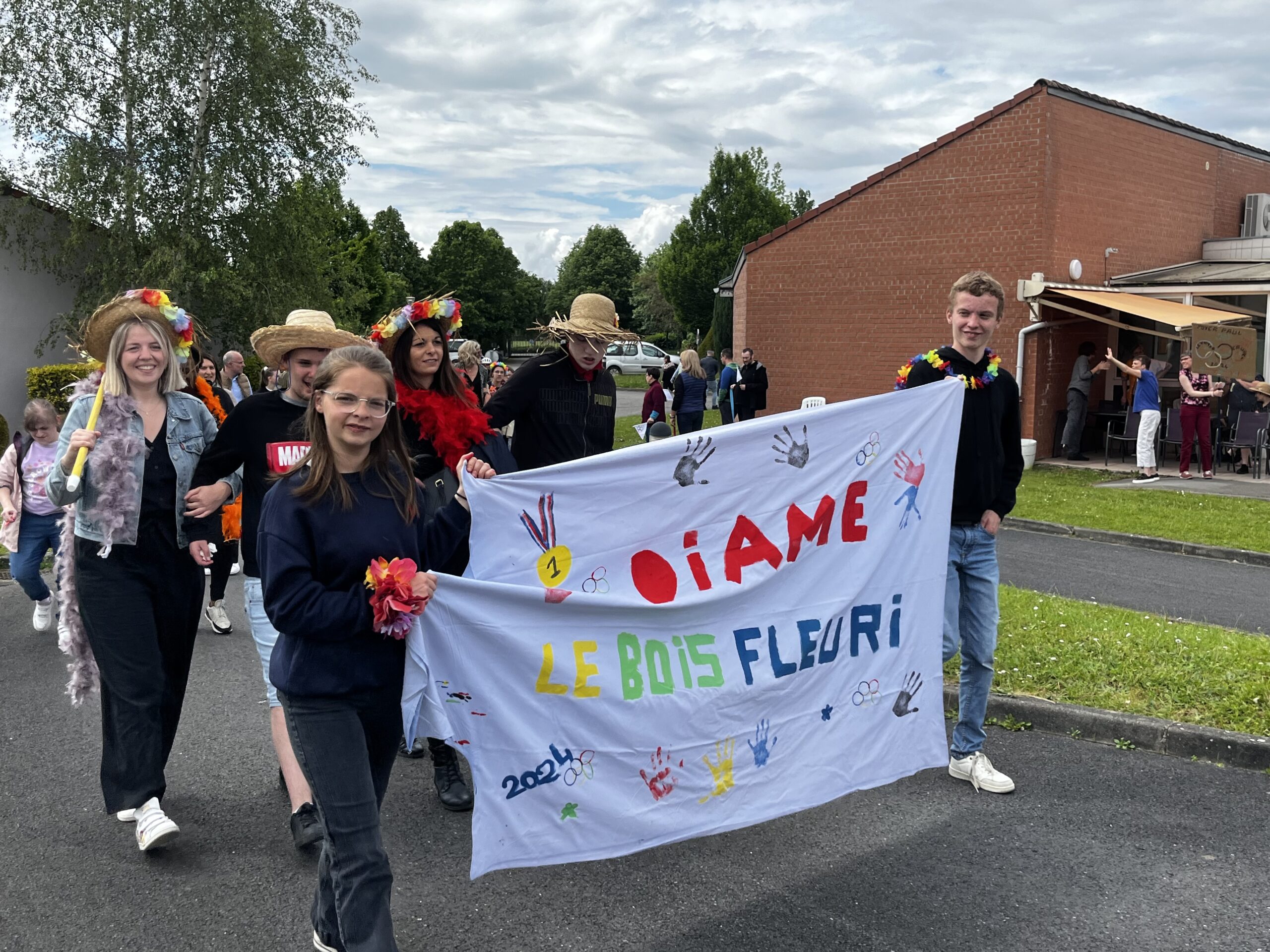 Cortège costumé portant une banderole peinte « Le Bois Fleuri 2024 », ornée d'anneaux olympiques et d'empreintes de mains.