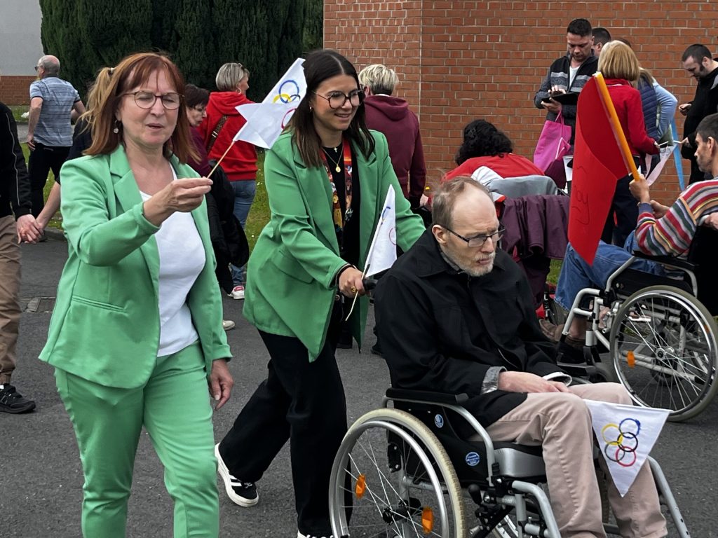 Cortège en extérieur : des participants agitent des fanions aux anneaux olympiques, dont un homme en fauteuil roulant.