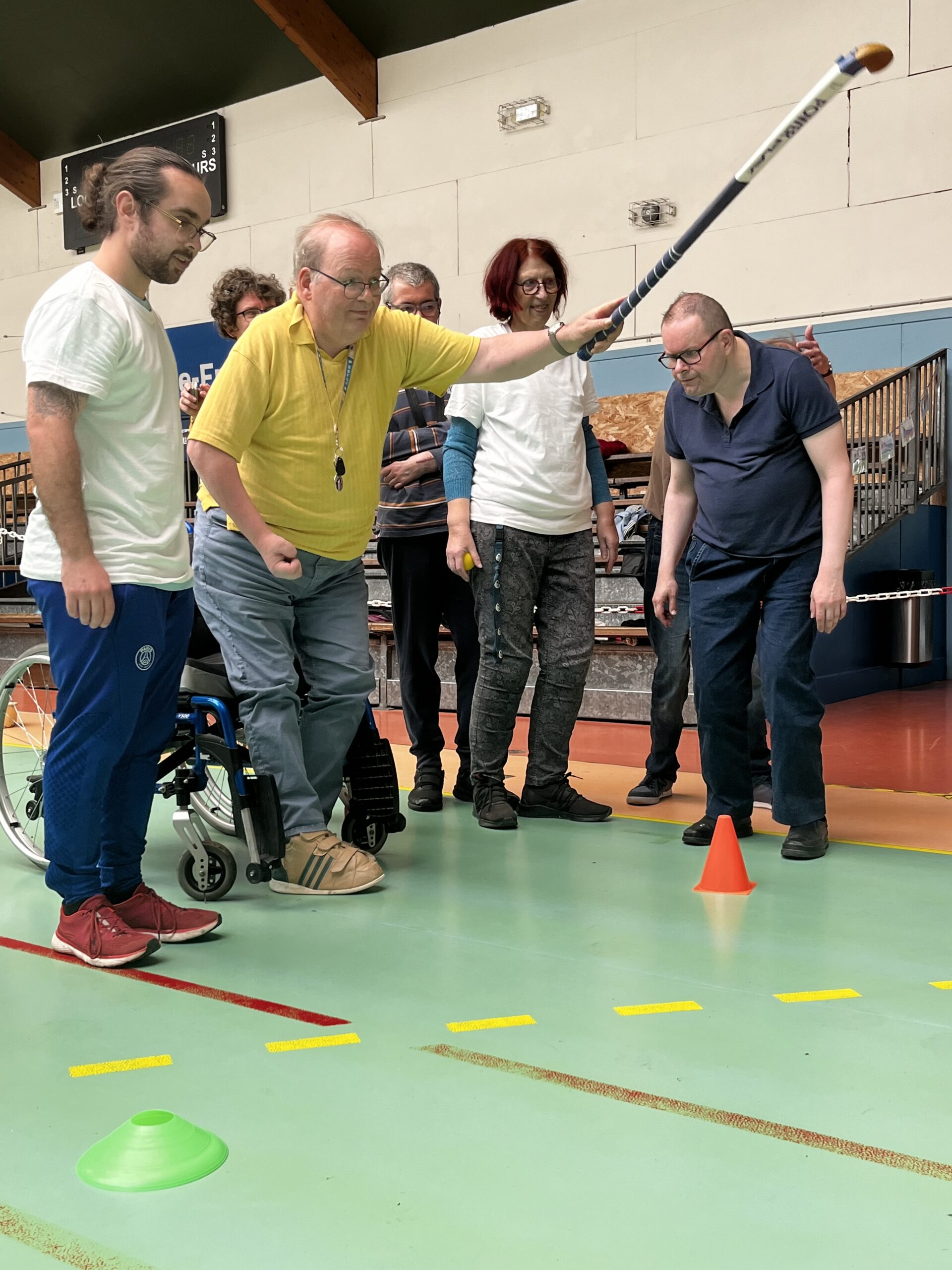 Dans un gymnase, un participant frappe une balle avec une crosse pendant un atelier de sport adapté, entouré du groupe.