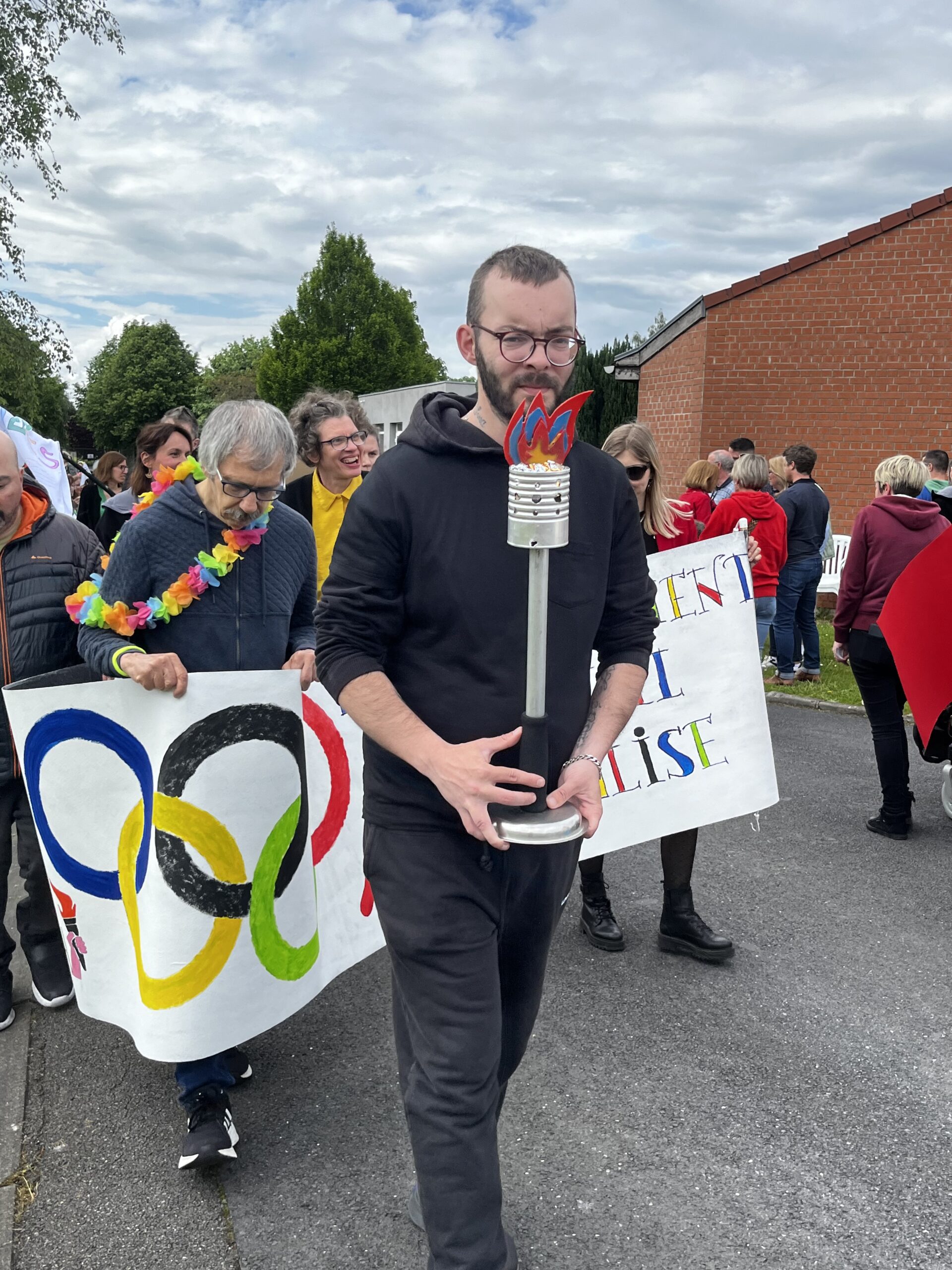 Un homme porte une torche olympique fabriquée à la main lors d'un défilé, derrière lui une banderole aux anneaux olympiques.