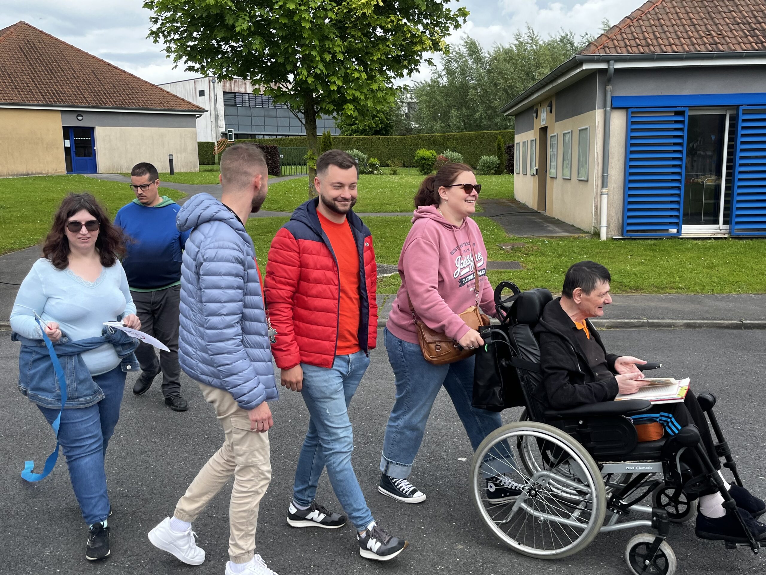Un groupe marche sur une allée, une femme pousse un homme en fauteuil roulant, un livre sur les genoux.