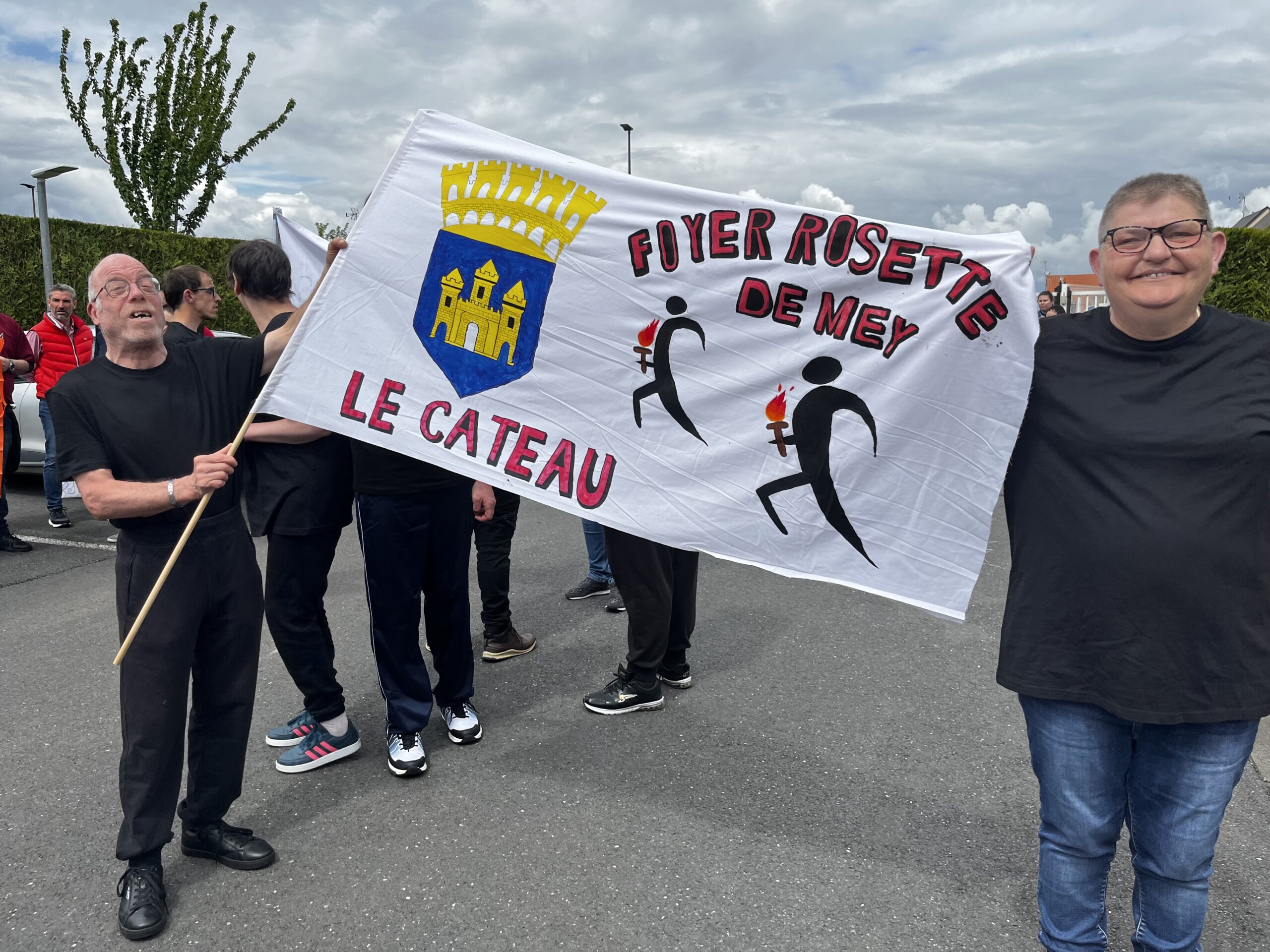 Deux personnes portent une banderole « Foyer Rosette de Mey - Le Cateau », ornée du blason de la ville et de porteurs de flamme.