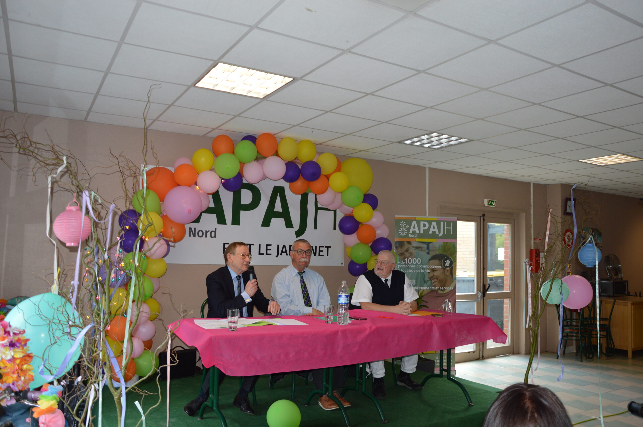 Quatre hommes à une table de tribune sous une arche de ballons, devant la banderole APAJH Nord ESAT Le Jardinet.