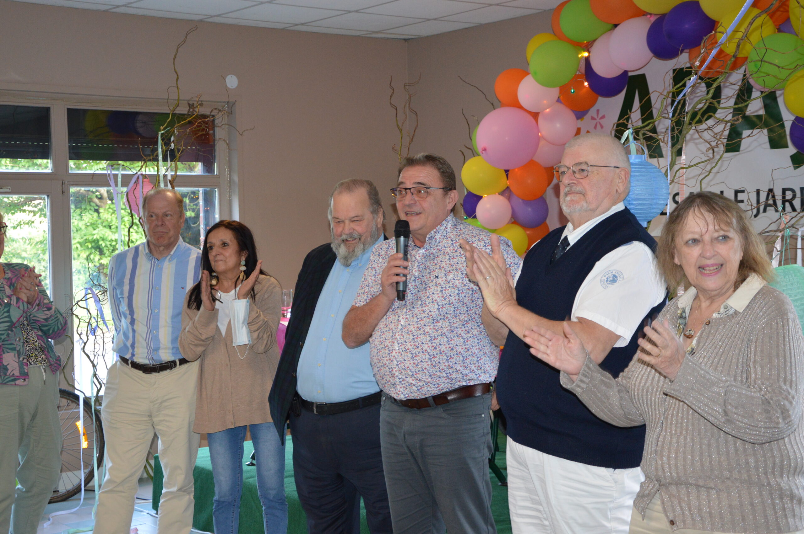 Une personne prend la parole au micro, entourée d'invités qui applaudissent, devant une arche de ballons colorés.
