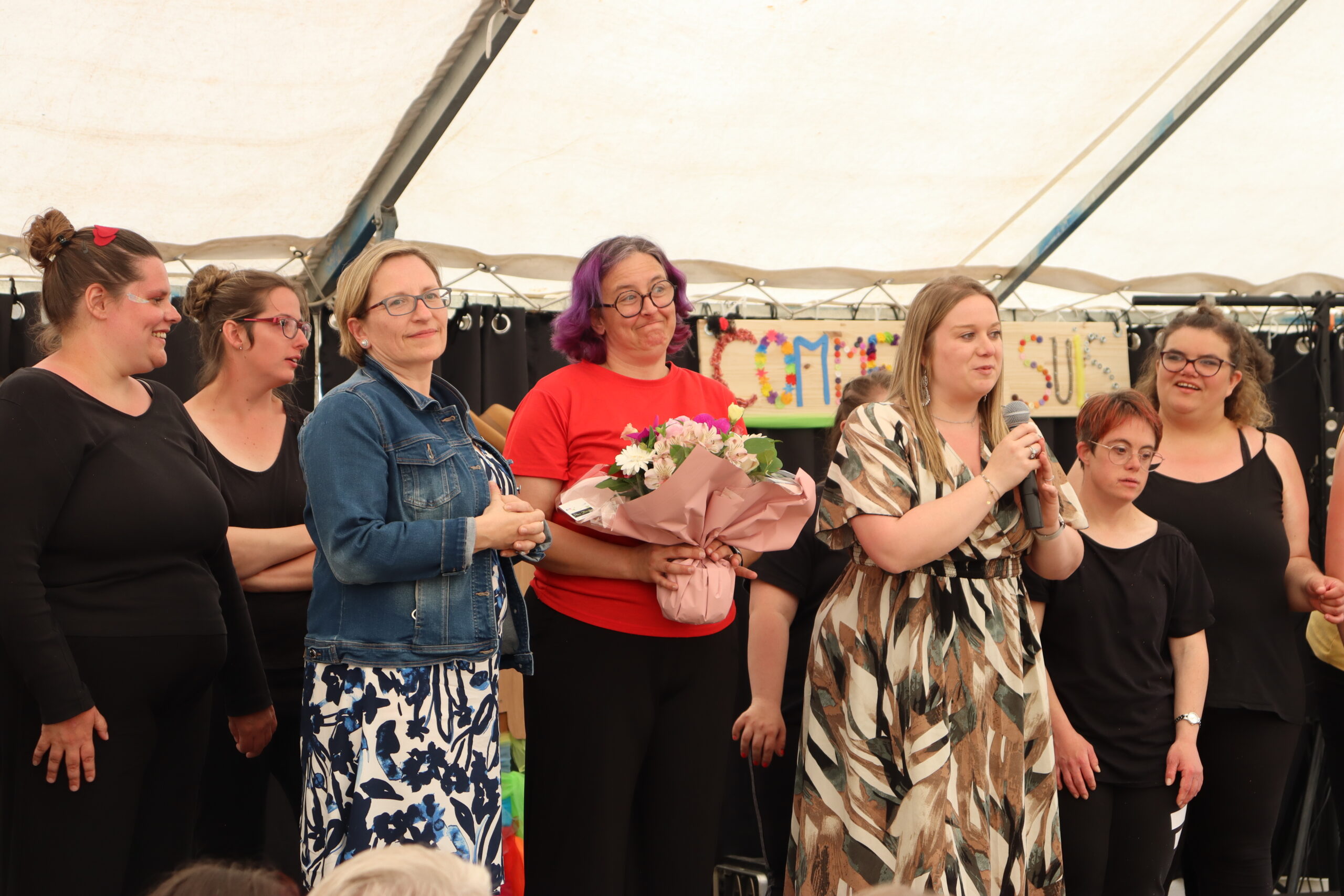 Sous un chapiteau, une prise de parole au micro à côté d'une femme tenant un bouquet, entourées de danseuses en noir.