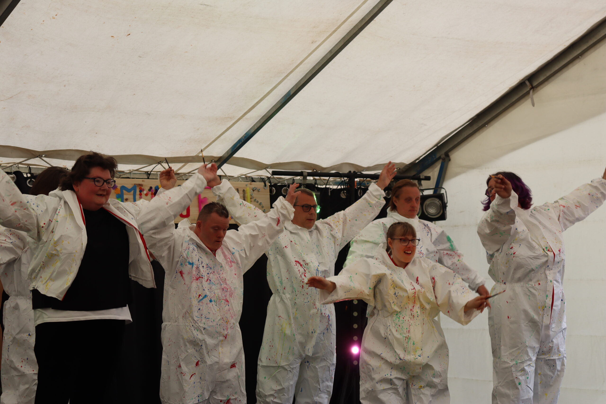 Danseurs en combinaisons blanches éclaboussées de peinture, pinceaux à la main, bras levés sur une scène sous chapiteau.