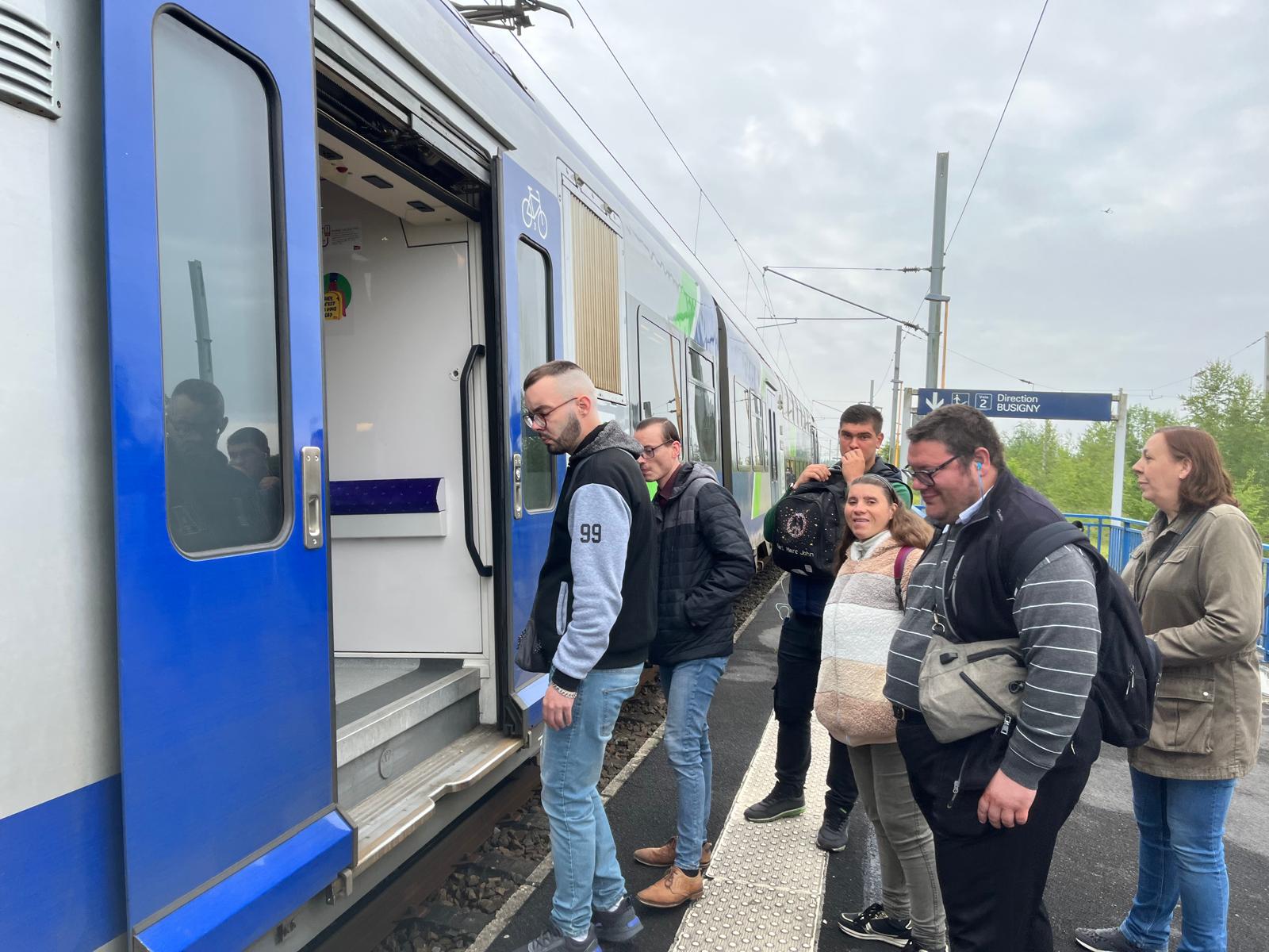 Sur un quai, un groupe d'adultes s'apprête à monter dans un TER ; le panneau indique « Voie 2, direction Busigny ».