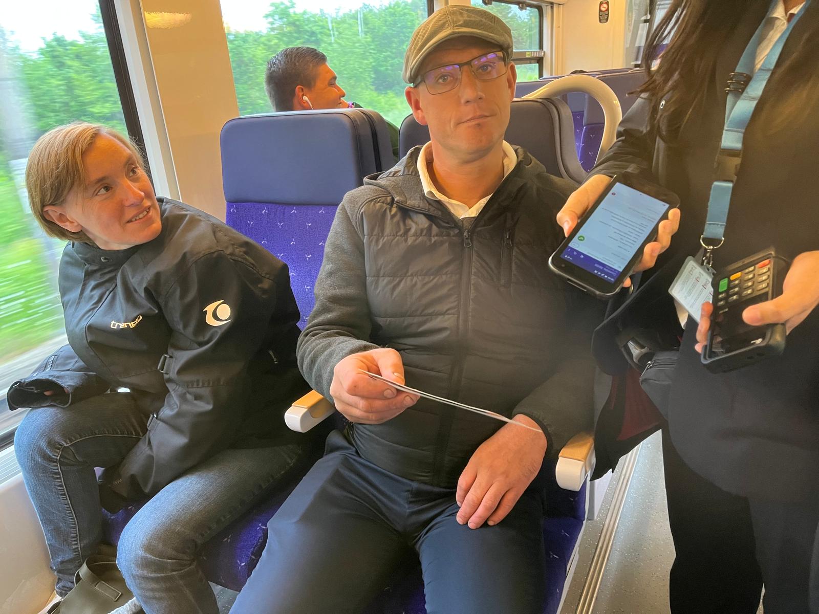 Dans un train, deux voyageurs assis pendant qu'une contrôleuse leur présente un smartphone et un terminal de paiement.