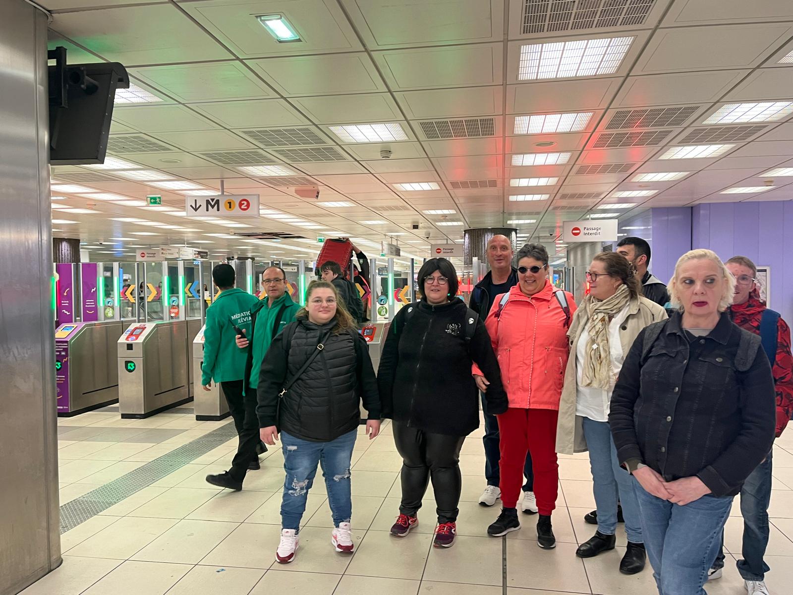 Un groupe d'adultes accompagné se tient devant les portiques d'une station de métro, sous les panneaux des lignes 1 et 2.