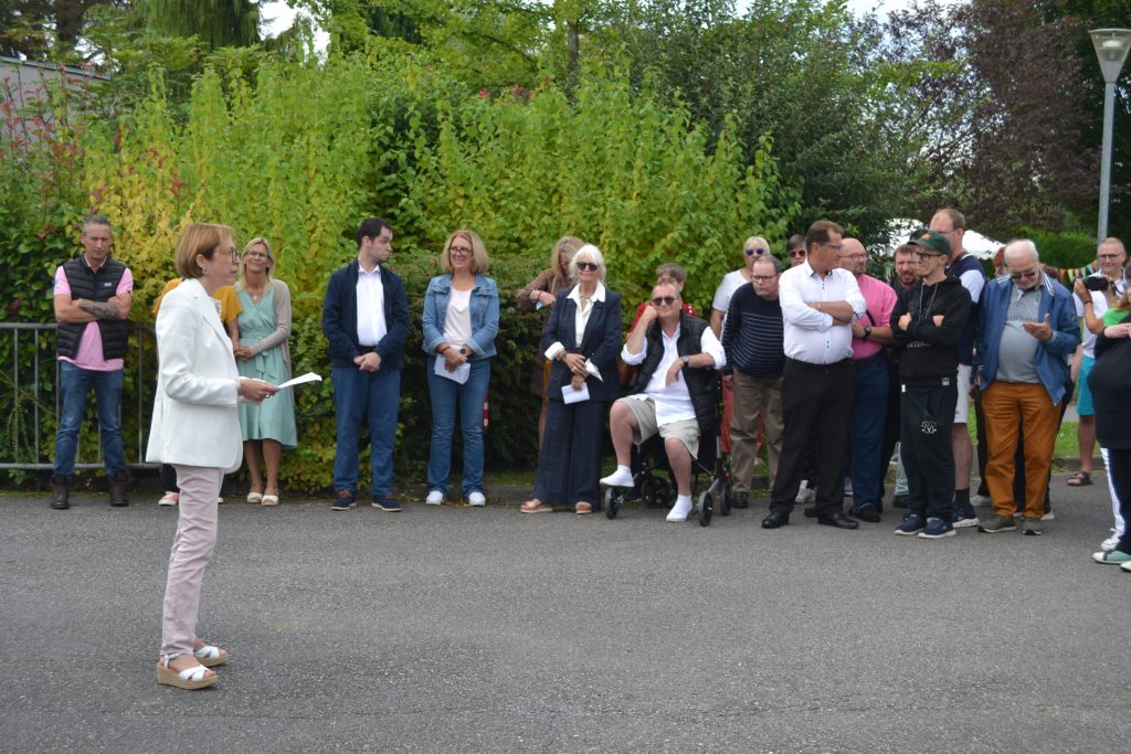 En plein air, une femme lit un discours devant un groupe debout le long d'une haie.