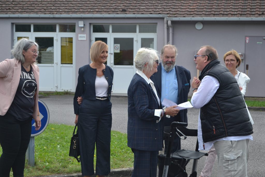 Un petit groupe d'adultes en tenue de ville discute devant l'entrée d'un bâtiment, papiers à la main.