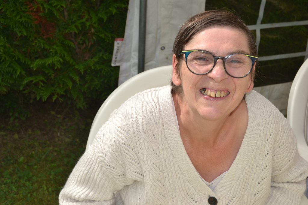 Portrait souriant d'une femme à lunettes, en gilet blanc, assise sous un chapiteau dans un jardin.
