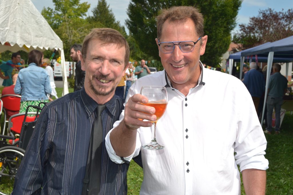 Deux hommes souriants lors d'une fête en plein air, l'un lève son verre ; barnums et guirlandes derrière eux.