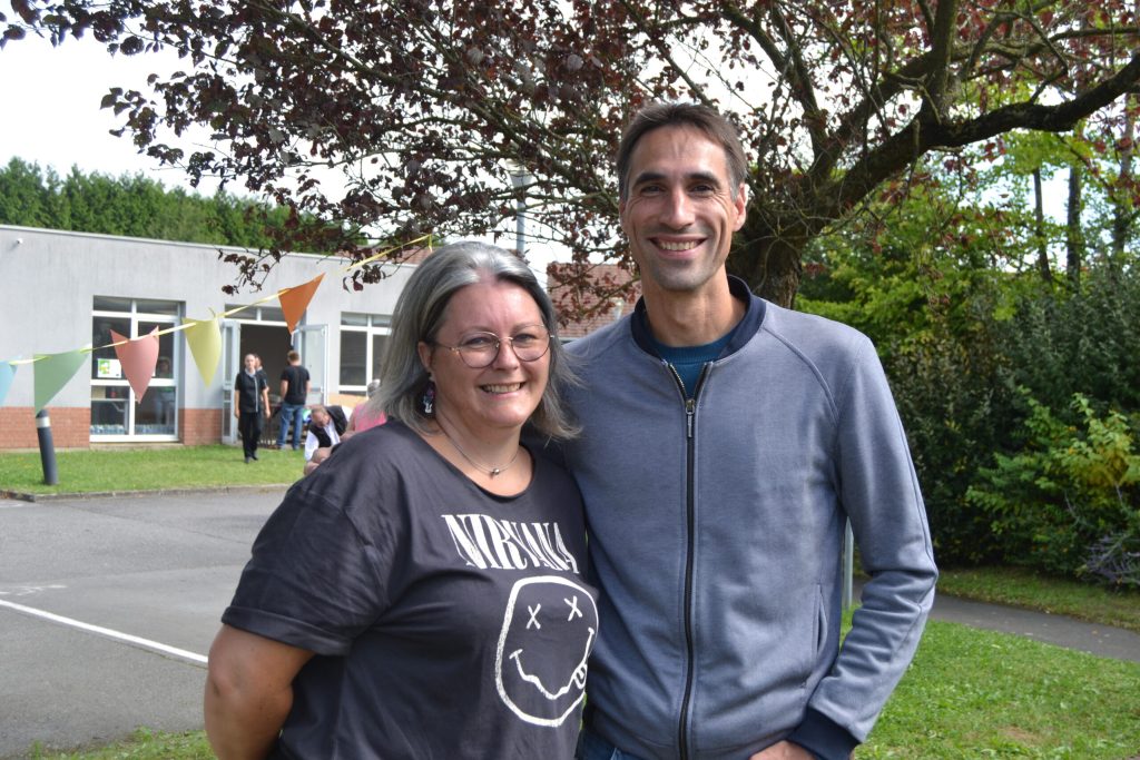 Deux personnes souriantes posent côte à côte dans un parc, devant un bâtiment décoré de fanions.