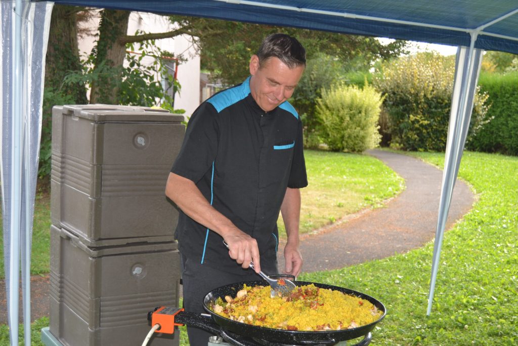 Sous une tonnelle, un cuisinier remue une grande paella dans le parc de l'établissement.