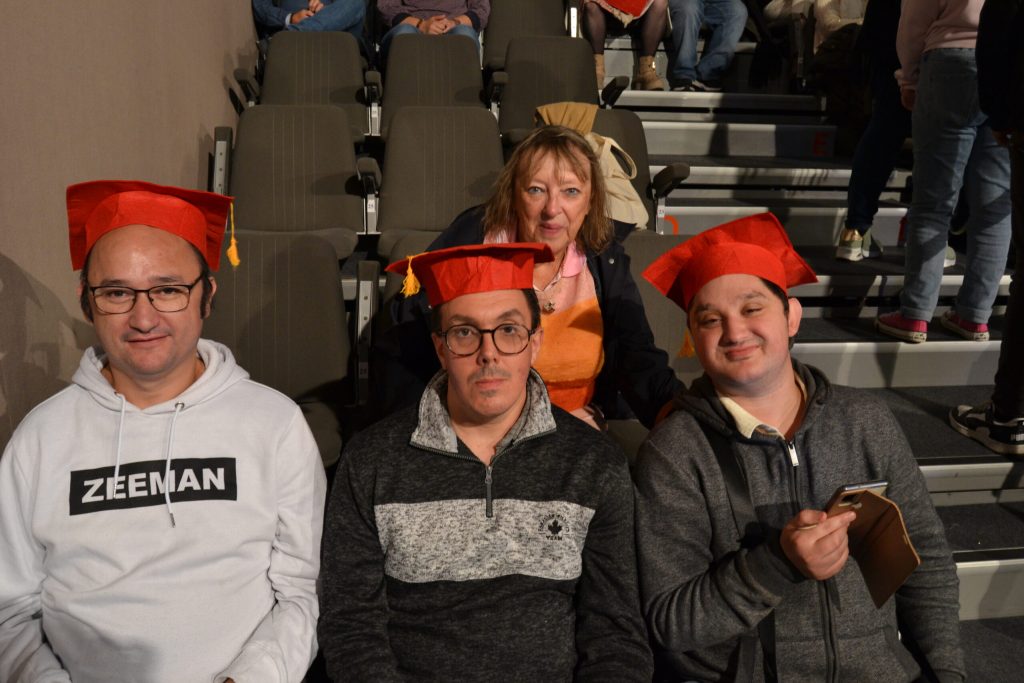 Trois hommes coiffés d'un chapeau de diplômé rouge, assis dans un gradin, lors d'une remise de diplômes.
